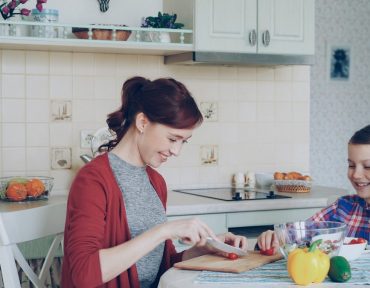 Happy young mother cooking vegetables cutting tomatoes and talking to her cheerful daughter in the kitchen at home. Family, cook, and people concept